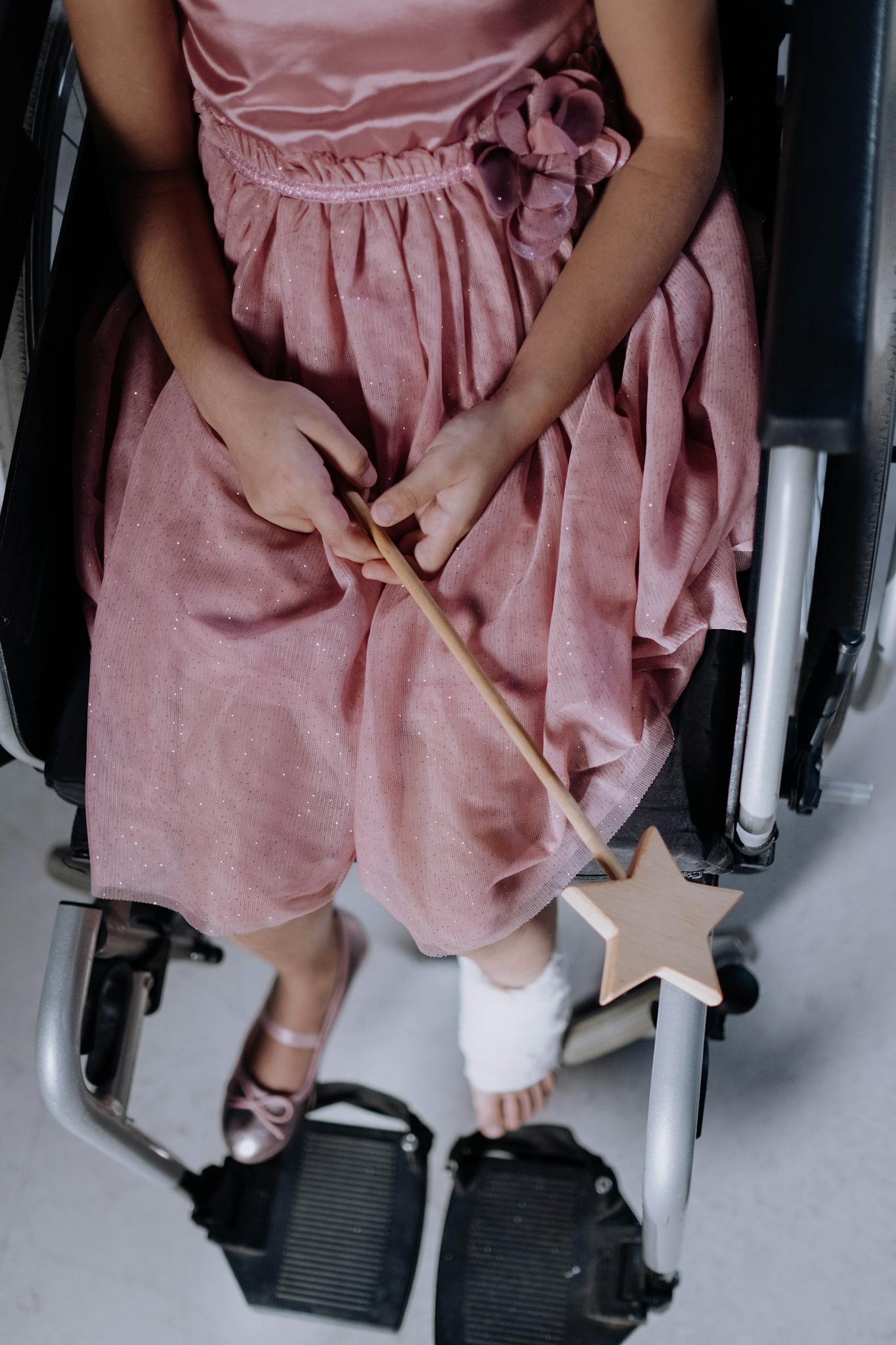 Girl in pink dress sitting in a wheelchair holding a magic wand, with an orthopedic cast on foot.